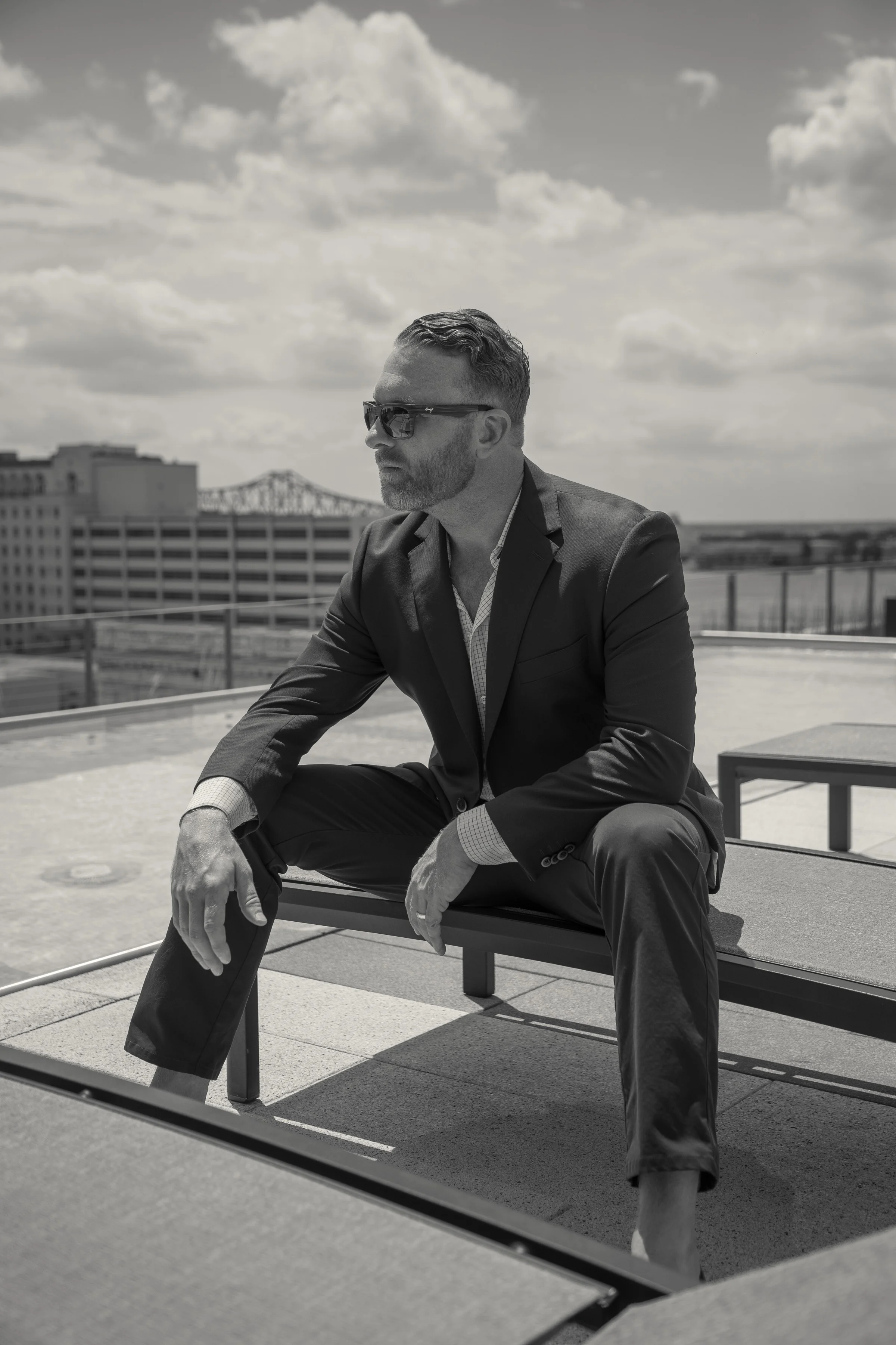 Dustin Alexander of Alexander Realty, LLC, seated on a downtown Baton Rouge rooftop in a dark suit, the Mississippi River bridge and skyline behind him.