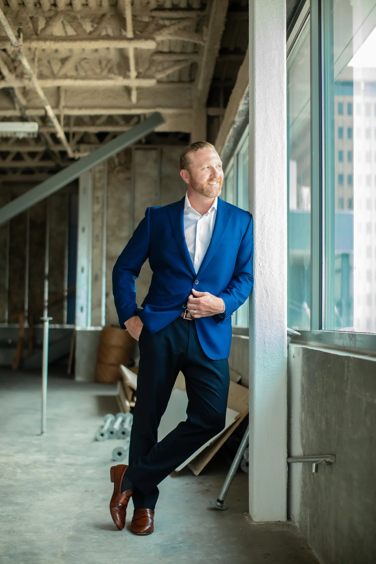 Dustin Alexander in a blue blazer, leaning against a column in a downtown high rise with the Baton Rouge skyline beyond the windows.